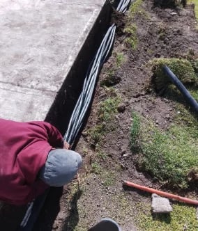 Person in maroon jacket working on underground electrical conduit installation along garden bed with moss and tools visible