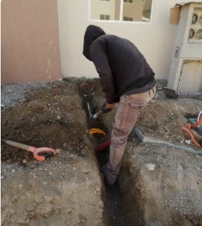 Person in black hoodie and torn pants digging in gravel pit near building entrance with tools visible