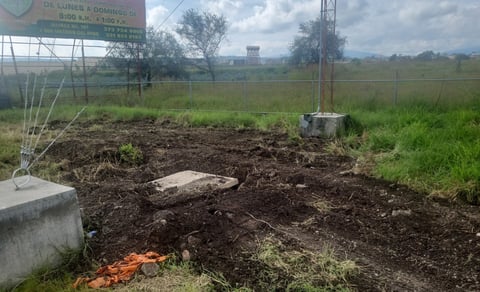 Mound of dark soil and rocks in rural field with green grass, metal fencing, and distant structures under cloudy sky