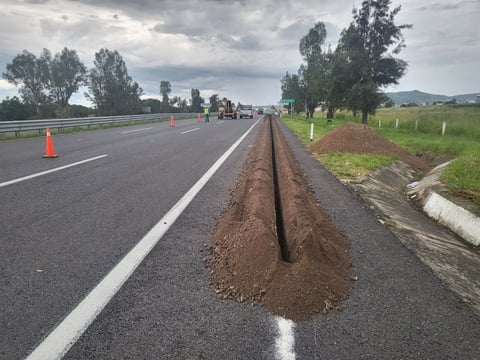 Highway construction site with drainage pipe installation, traffic cones, and rural landscape with trees under cloudy sky