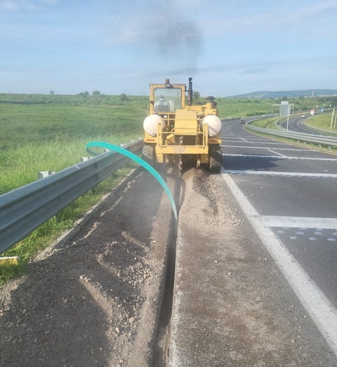 Yellow road maintenance truck spraying tar on highway shoulder with green fields and distant hills in background