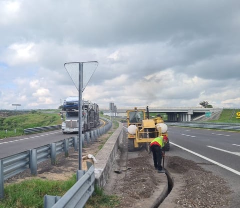 Yellow construction vehicles working on highway shoulder with truck and traffic sign visible