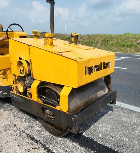 Yellow Ingersoll Rand road roller compactor on asphalt pavement with vegetation and utility pole in background
